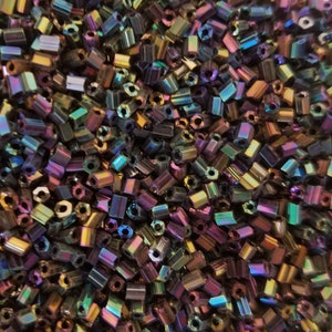 May include: A close-up shot of a pile of iridescent, rectangular beads. The beads have a metallic sheen, reflecting colors like purple, green, and gold. The beads are small and uniform in size, suggesting they are for jewelry making or crafts.