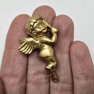 Può includere: Statuetta di cherubino dorata con ali dettagliate e posa giocosa. Il cherubino tiene in mano un oggetto, forse un arco o una freccia. La statuetta è posta su uno sfondo neutro.