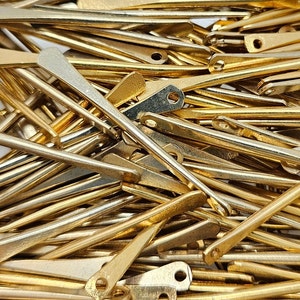 May include: A pile of gold-colored metal pieces, each with a pointed end and a small hole at the other end. The pieces are arranged in a random pattern, creating a textured and metallic background.