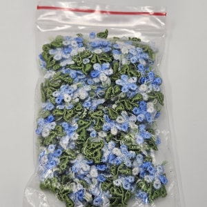 Könnte beinhalten: Ein klarer Plastikbeutel gefüllt mit floralen Verzierungen. Die Verzierungen sind eine Mischung aus Blau, Weiß und Grün, die kleinen Blumen und Blättern ähneln. Der Beutel ist mit einem roten Reißverschluss verschlossen.