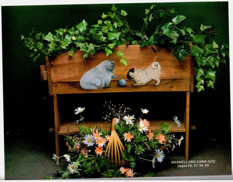 May include: A wooden shelf unit with two levels, decorated with painted pug and cat figures. The top shelf has a blue ball between the figures. The bottom shelf holds a basket of flowers. The text "MAXWELL AND KIMMI SOO pages 56, 57, 58, 59" is visible.