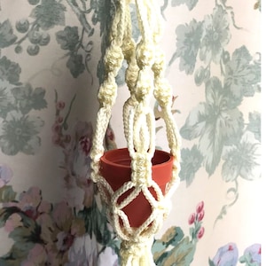May include: A white macrame plant hanger with a small terracotta pot. The hanger is attached to a metal ring and has a tassel at the bottom.