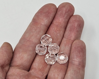 May include: A hand holding five clear, faceted beads. The beads have a multi-faceted design, catching the light. The beads are a pale, translucent color, and appear to be made of glass or crystal. The background is a plain white surface.