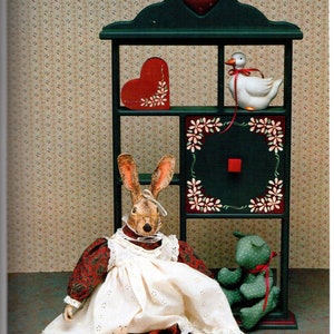 May include: A dark green shelf with a heart-shaped top, holding a white duck figurine, a red heart, and a square with floral accents. A rabbit doll in a white dress and a green teddy bear are in front of the shelf. The text "WHAT NOT SHELF AND HEART" is visible.