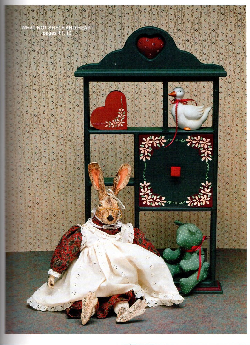 May include: A dark green shelf with a heart-shaped top, holding a white duck figurine, a red heart, and a square with floral accents. A rabbit doll in a white dress and a green teddy bear are in front of the shelf. The text "WHAT NOT SHELF AND HEART" is visible.