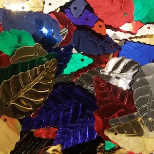 May include: A close-up shot of metallic leaf-shaped confetti in various colors, including gold, silver, red, blue, and green. Each leaf has a small hole at the top. The leaves are scattered, creating a vibrant and festive display.