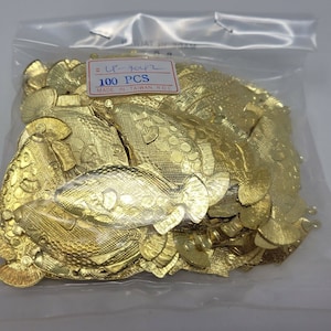 May include: A clear plastic bag containing 100 gold-colored metal fish-shaped charms. Each flat charm features a textured scale pattern and a small loop at the top. A white label on the bag displays the text 100 PCS and MADE IN TAIWAN R.O.C.
