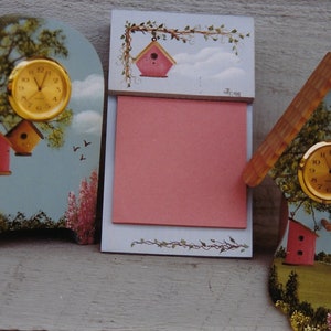 Könnte beinhalten: Drei dekorative Holzartikel: eine Uhr, ein Notizblock und eine hausförmige Uhr. Die Uhr hat ein goldenes Zifferblatt und ist vor einem bemalten Hintergrund mit einem Vogelhaus, Bäumen und Himmel angebracht. Der Notizblock hat einen rosafarbenen Block.