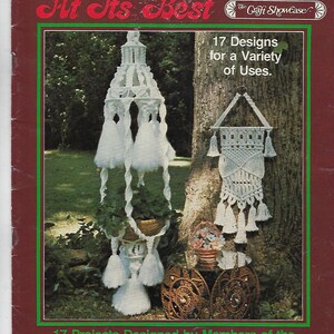 Puede incluir: Una portada de libro con el título "Macrame At Its Best" que presenta una planta colgante de macramé blanca y una pared de macramé blanca. La portada del libro es marrón con bordes verdes y texto en blanco y rojo. El texto de la portada dice "17 Designs for a Variety of Uses." y "17 Projects Designed by Members of the Craft Showcase Team, Featuring Projects Created Especially for You."