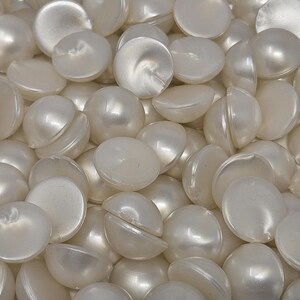 May include: A close-up shot of a pile of white, half-sphere shaped beads with a pearlescent sheen. Some beads are whole, while others are split in half, revealing a hollow interior. The beads are scattered, creating a textured surface.