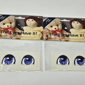 May include: Two sets of doll eye patches. Each set includes two embroidered blue eyes with white highlights and black outlines. The packaging features images of dolls and the text "The Eyes Have It!"