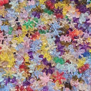 May include: A large collection of small, translucent, multi-colored plastic beads shaped like six-petaled flowers. The beads are scattered together in a dense pile, featuring vibrant shades of red, yellow, blue, green, purple, and pink.