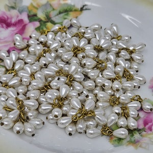 May include: A pile of white pearl teardrop beads with gold-toned metal loops. The beads are arranged in a loose cluster on a white surface with a floral pattern.