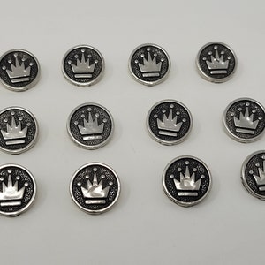 Pode incluir: Doze botões de metal prateados com um design de coroa. Cada botão é redondo e tem um design de coroa em relevo no centro.