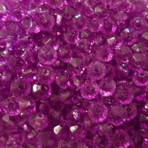 May include: A collection of small, translucent, vibrant purple beads with a faceted surface. Each bead features a central hole, creating a uniform appearance across the pile. The light reflects off the angled surfaces of the beads, highlighting their deep magenta hue and circular shape throughout the entire frame.