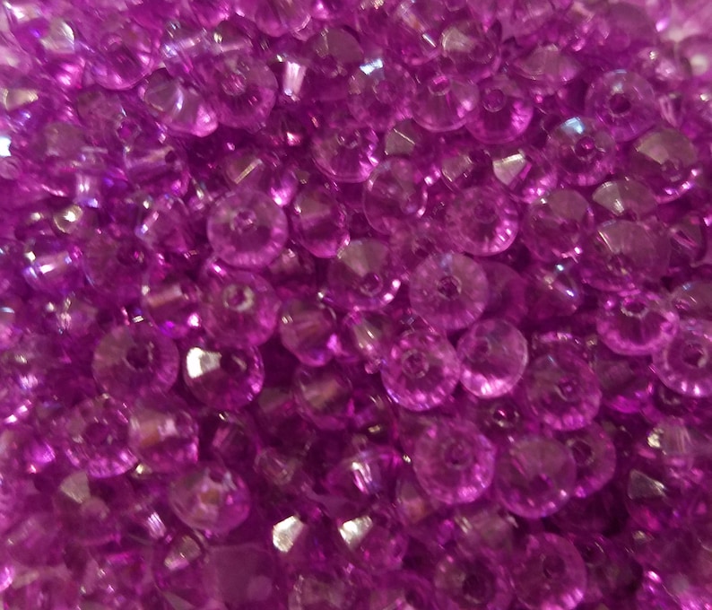 May include: A collection of small, translucent, vibrant purple beads with a faceted surface. Each bead features a central hole, creating a uniform appearance across the pile. The light reflects off the angled surfaces of the beads, highlighting their deep magenta hue and circular shape throughout the entire frame.