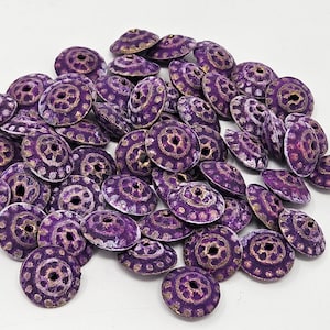 May include: A collection of purple, disc-shaped beads with a central hole and a gold-colored, dotted pattern. The beads have a metallic finish and are scattered on a white surface. These beads are suitable for jewelry making and crafts.