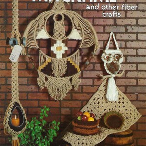 May include: A book cover with the title "Macrame' and other fiber crafts" in white text on a brick background. The cover features a variety of macrame projects, including a large wall hanging, a small owl-shaped hanging, and a crocheted throw pillow.