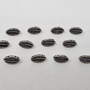 May include: A collection of 12 silver-toned metal buttons shaped like feathers. The buttons are arranged in a semi-circle on a white background.
