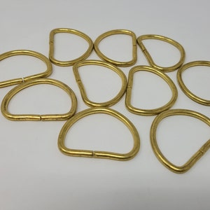 May include: Ten gold-colored metal D-rings, also known as split rings, are arranged in a circle on a white background. These rings are commonly used for attaching items to keychains, bags, and other accessories.