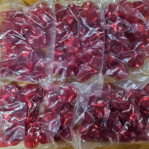 May include: Several clear plastic bags filled with translucent red heart-shaped objects. The bags are sealed and arranged on a wooden surface. The hearts appear to be made of a shiny, smooth material.