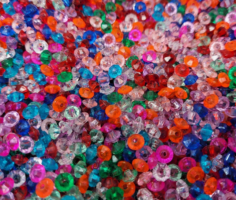 May include: A large collection of small, faceted plastic beads in a vibrant mix of translucent colors including red, orange, blue, green, pink, and clear. Each bead features a central hole and a multi-faceted surface that reflects light.