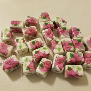 May include: A collection of white ceramic square beads with pink floral designs. The beads have a hole through the center for stringing.