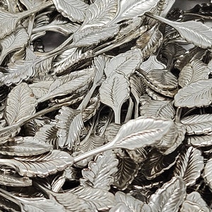 May include: A pile of silver-colored metal leaf-shaped charms. The charms are all the same size and shape, and they are arranged in a random pattern.