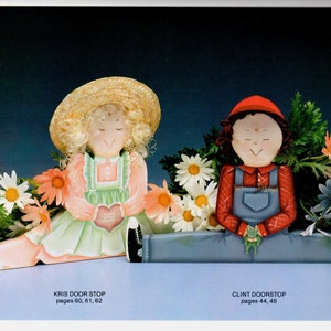 May include: Two painted wooden doorstops, one depicting a girl in a straw hat and the other a boy in a red cap. Both are surrounded by painted flowers. The girl holds a heart. Text at the bottom reads "KRIS DOOR STOP" and "CLINT DOORSTOP".