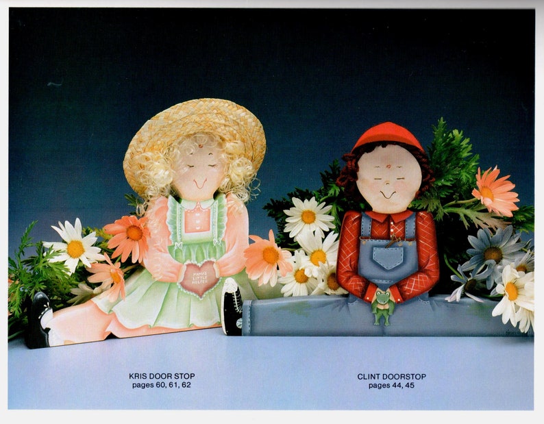May include: Two painted wooden doorstops, one depicting a girl in a straw hat and the other a boy in a red cap. Both are surrounded by painted flowers. The girl holds a heart. Text at the bottom reads "KRIS DOOR STOP" and "CLINT DOORSTOP".