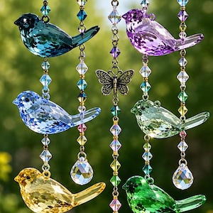 May include: A hanging mobile featuring seven faceted glass birds in blue, purple, light blue, light green, yellow, green, and pink. The birds are suspended from a black metal ring by chains adorned with colorful crystal beads and a central butterfly.