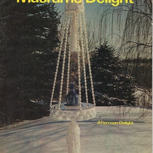 Peut inclure: Un pot de fleurs suspendu en macramé blanc avec un pompon blanc et un vase en céramique bleu et blanc à l'intérieur. Le pot est suspendu à un cordon en macramé blanc et est suspendu au-dessus d'une table en macramé blanc avec un plateau rond et un pompon blanc. Le texte "Macrame Delight" est en lettres jaunes en haut de l'image. Le texte "Book One" est en lettres jaunes au-dessus du texte "Macrame Delight". Le texte "Afternoon Delight" est en lettres jaunes sous la table. Le texte "by Anne" est en lettres jaunes en bas à droite de l'image.