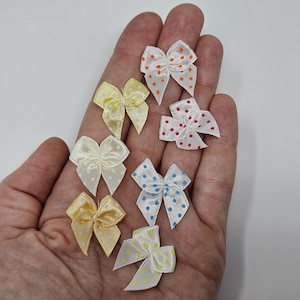 Könnte beinhalten: Eine Sammlung kleiner Satin-Schleifen in verschiedenen Farben, darunter Gelb, Weiß, Orange und Blau. Jede Schleife ist mit Polka-Dots verziert. Die Schleifen sind auf hellem Hintergrund angeordnet.