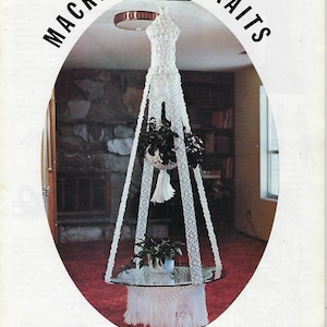 May include: A white macrame hanging plant holder with a glass table top. The macrame is in the shape of a bell and has a fringe at the bottom. The plant holder is hanging from the ceiling in a room with a fireplace and a bookcase. The image is from a book titled "Macrame Portraits" Book 3, 150. The caption below the image reads "Glass Slipper" by Vickie.