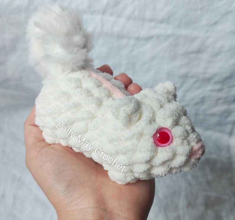 Puede incluir: Un animal de peluche tejido a ganchillo en color blanco con una cola esponjosa, ojos de pl&aacute;stico rosa y una peque&ntilde;a nariz rosa. El texto Lolly May Crochet est&aacute; impreso en el costado del cuerpo de hilo blanco. Hecho de hilo blanco grueso y suave.