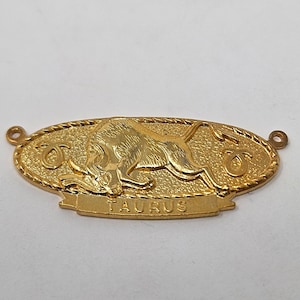 May include: Gold-colored metal charm with a detailed image of a bull and the word "TAURUS" inscribed on the bottom. The charm has two small loops on either side for attaching to a necklace or bracelet.
