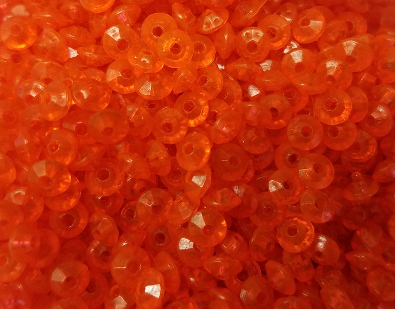 May include: A collection of small, round, translucent orange beads with a faceted surface and a central hole. Each bead features a circular shape with multiple angled cuts that reflect light, creating a bright, uniform orange appearance throughout.