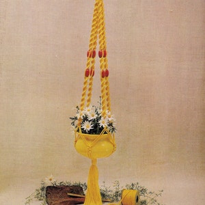 May include: A yellow macrame plant hanger with red beads, holding a yellow ceramic pot with white flowers. The hanger is hanging from a ceiling hook and has a long tassel. There is a ball of yellow yarn and a piece of wood on the floor.