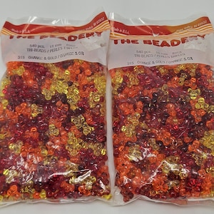 Op de afbeelding: Twee doorzichtige plastic zakken gevuld met oranje, goudkleurige en rode tri-beads. De zakken hebben een rode en oranje koptekst met de tekst "THE BEADERY". Elke zak bevat 540 kralen van 11 mm. De kralen zijn gemarkeerd met "ORANGE & GOLD".