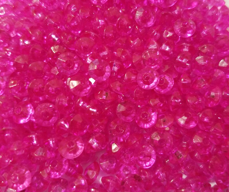 May include: A collection of small, round, translucent hot pink beads with a faceted surface texture. Each bead features a central hole for threading, creating a uniform appearance of bright, reflective pink circular shapes packed together.