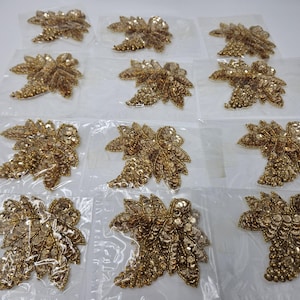 以下が含まれることがあります： 白い背景に、金色の葉の形をした装飾品が12個、並べて置かれています。装飾品には、スパンコールが施されています。