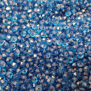 May include: A large collection of small, transparent, bright blue faceted beads. Each bead features a multi-sided, geometric cut that reflects light, creating a sparkling appearance across the dense, uniform pile of loose craft components.