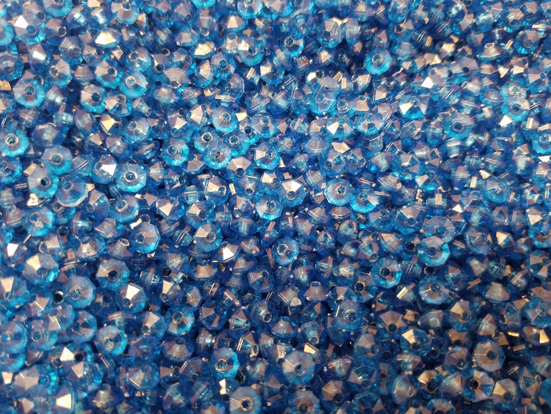 May include: A large collection of small, transparent, bright blue faceted beads. Each bead features a multi-sided, geometric cut that reflects light, creating a sparkling appearance across the dense, uniform pile of loose craft components.