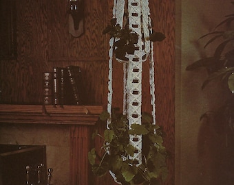 May include: A white macrame plant hanger with a woven design hangs from the ceiling. The hanger is filled with green plants and has a long tassel at the bottom. The hanger is in front of a wooden wall with a fireplace below.