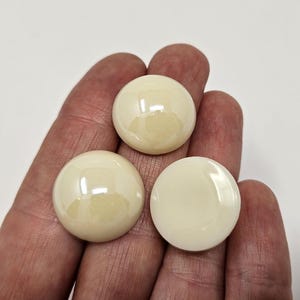 以下が含まれることがあります： 光沢のあるオフホワイトの丸いカボションが3つ、手に持たれています。カボションはわずかにドーム型で、光を反射してハイライトを作り出しています。背景は無地の白です。