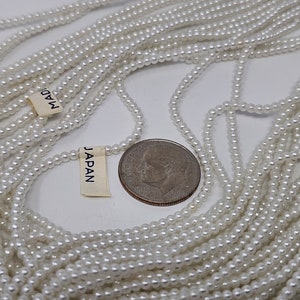 May include: A close-up of a large quantity of white pearl beads, with a US quarter for scale. The beads are strung on a thread and are arranged in a pile. The beads are labeled "MADE IN JAPAN".