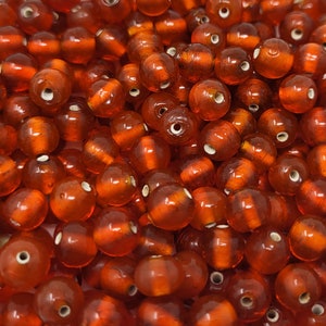 May include: A close-up image of a large quantity of small, round, translucent amber-coloured glass beads. The beads are scattered randomly and appear to be made of a smooth, shiny material.