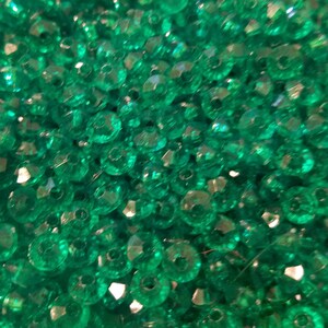 May include: A collection of small, round, translucent emerald green beads with faceted surfaces. Each bead features a central hole for threading. The beads are clustered together, creating a dense, reflective, and vibrant green textured surface.