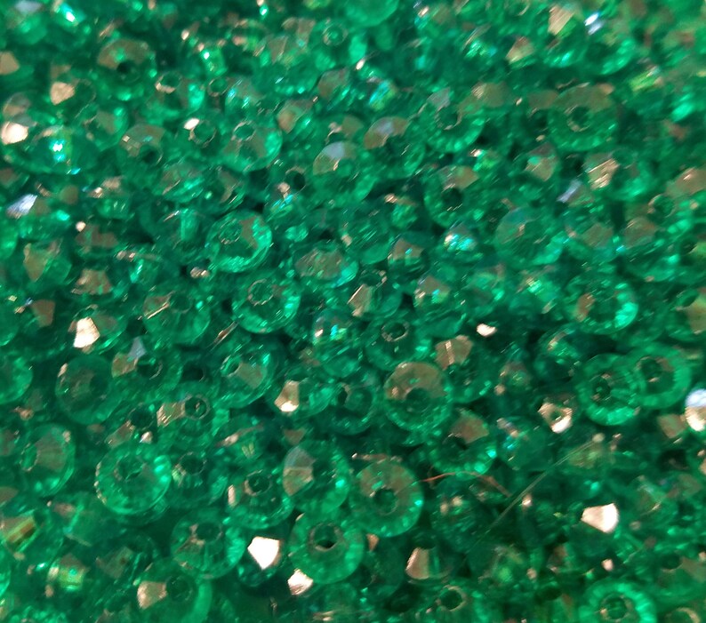 May include: A collection of small, round, translucent emerald green beads with faceted surfaces. Each bead features a central hole for threading. The beads are clustered together, creating a dense, reflective, and vibrant green textured surface.