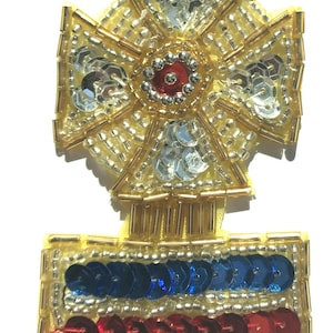 May include: A gold and silver beaded cross with a red and silver centre, and a blue, white and red beaded rectangle below it.